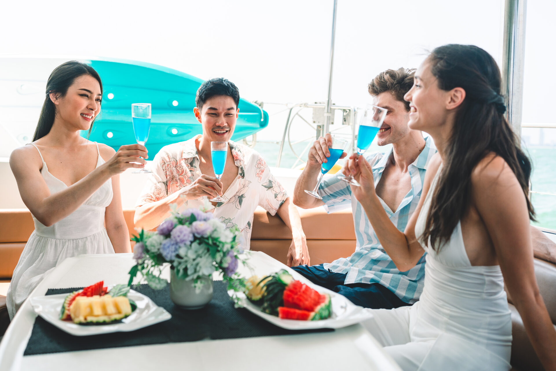 Group of diverse friends drink champagne while having a party in yacht. Attractive young men and women hanging out, celebrating holiday vacation trip while catamaran boat sailing during summer sunset.